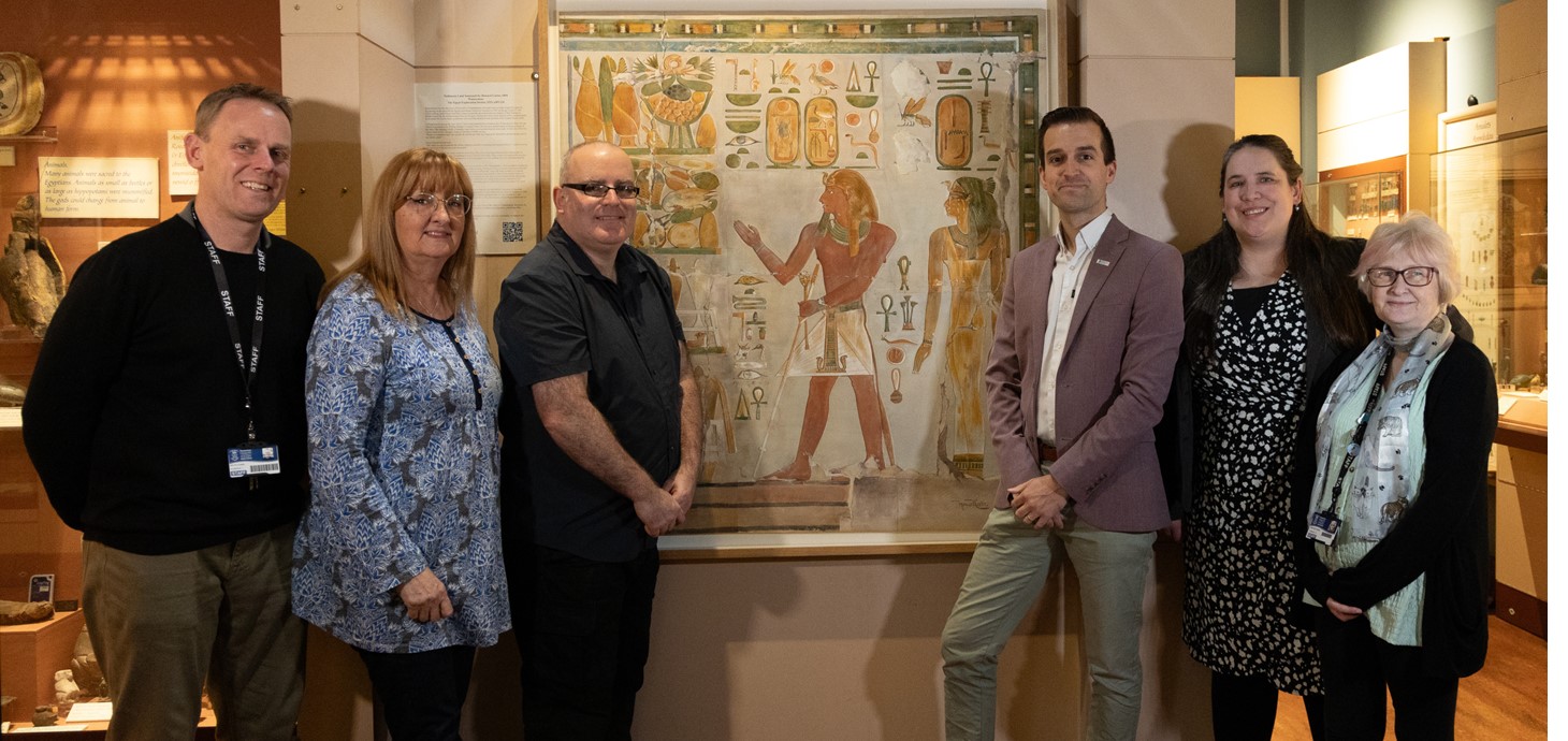 Group of six people standing either side of a large Egyptian painting in a gallery